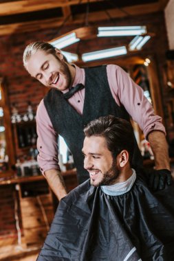 handsome barber in latex gloves putting hand on shoulder of happy man in barbershop  clipart