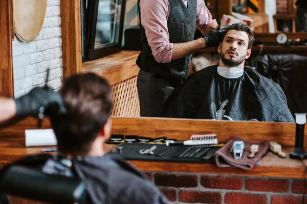 selective focus of barber styling hair of man near mirror in barbershop
