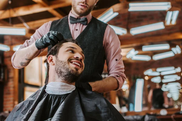 bearded barber in black latex gloves touching hair of happy man in barbershop