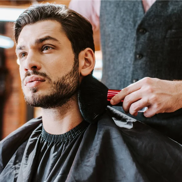 selective focus of barber holding cosmetic brush near face of handsome bearded man