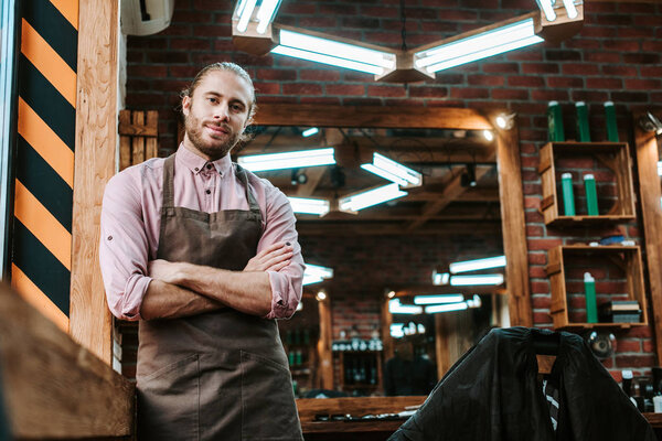 selective focus of handsome barber in apron standing with crossed arms and looking at camera