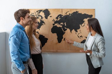 Gülümseyen seyahat acentesi, iki turistin yanındaki haritayı işaret ediyor.