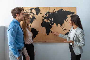 smiling travel agent pointing with finger at map on wall near couple of tourists clipart