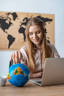 smiling travel agent touching globe while sitting at workplace clipart