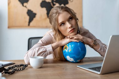 tired travel agent leaning on globe while sitting at workplace  clipart