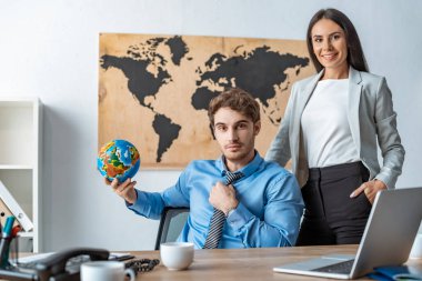 attractive travel agent standing with hand in pocket near colleague holding globe clipart