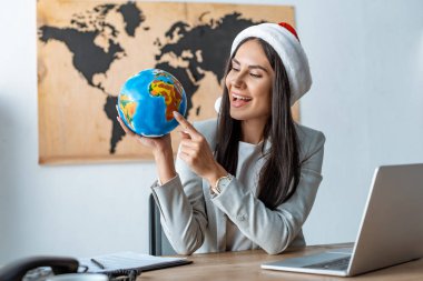 Noel Baba şapkalı gülümseyen seyahat acentesi işyerinde otururken dünyayı işaret ediyor.