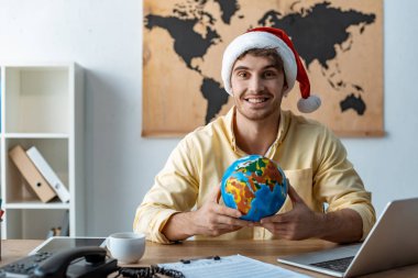 Noel Baba şapkalı yakışıklı seyahat acentesi kameraya gülümserken dünyayı tutuyor.