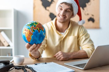 Noel Baba şapkalı neşeli seyahat acentesinin seçici odağı iş yerinde otururken dünyayı elinde tutuyor.