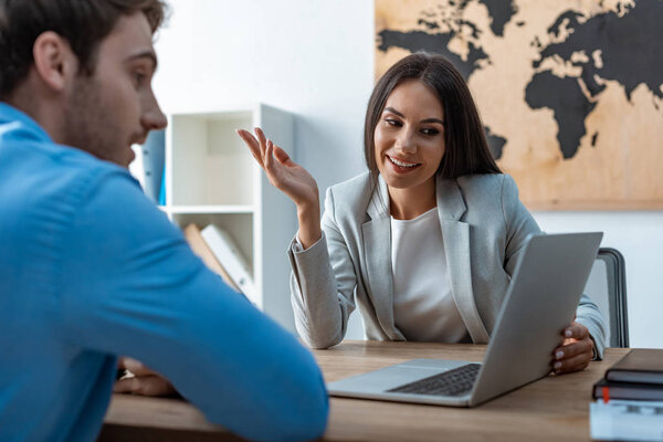 smiling travel agent looking at laptop screen while talking to client in office
