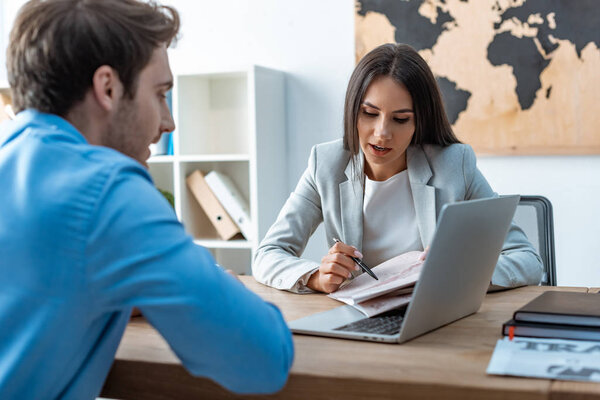 attractive travel agent pointing with pen at map while talking to client