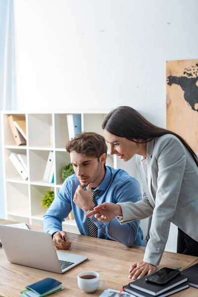 attractive travel agent pointing with finger at laptop near colleague sitting at workplace
