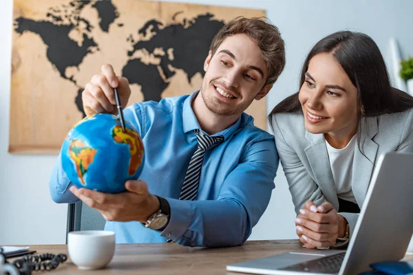 attractive travel agent standing near smiling colleague pointing with pen at globe
