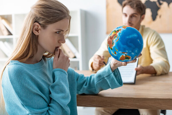 selective focus of thoughtful young woman looking at globe while sitting near travel agent
