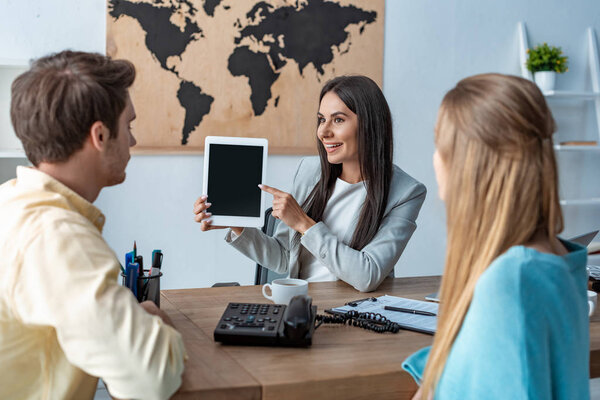 smiling travel agent pointing with finger and digital tablet near couple of tourists