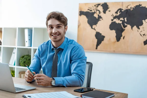 handsome travel agent smiling at camera while sitting at workplace