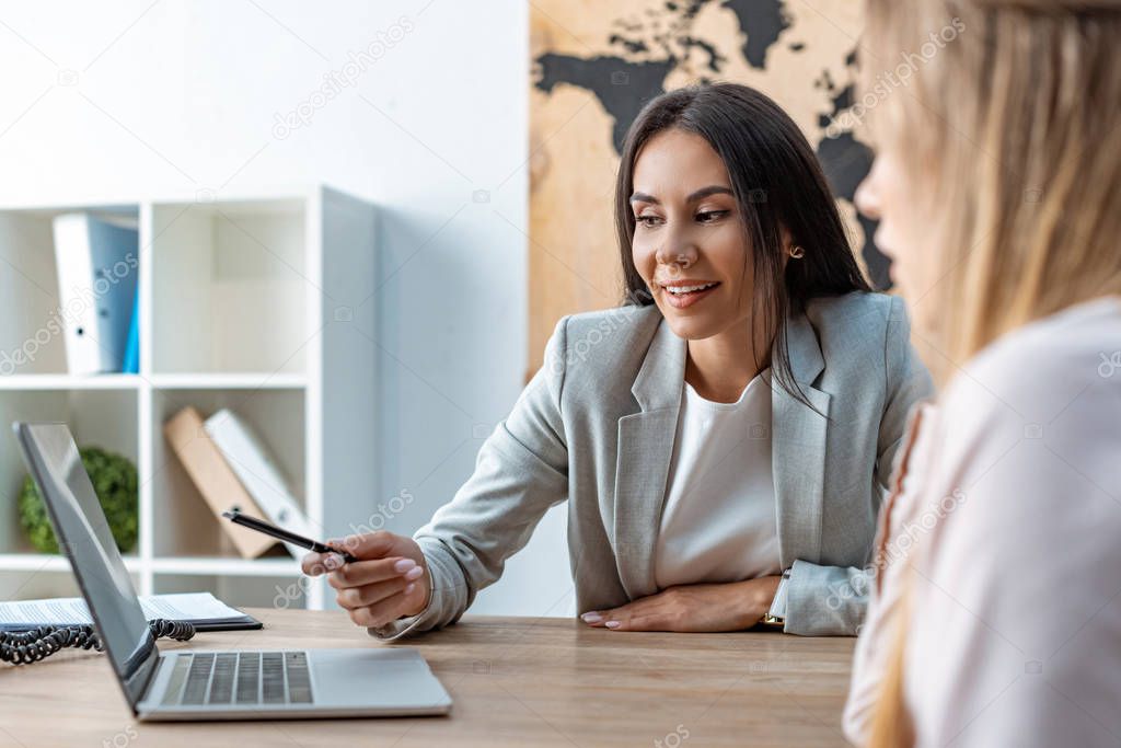 selective focus of positive travel agent pointing with pen at laptop screen near client