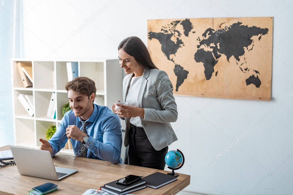 smiling travel agent pointing with hand at computer monitor near attractive colleague holding cup of coffee