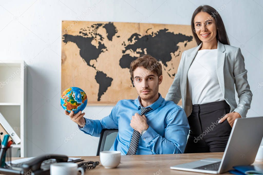 attractive travel agent standing with hand in pocket near colleague holding globe