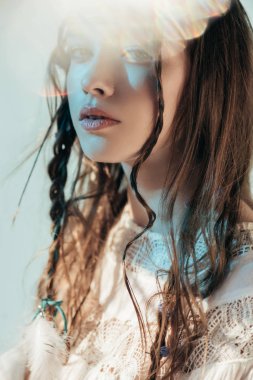 Saç stili örülü yumuşak bir kız. Beyaz boho elbisesiyle poz veriyor. Gri renkli lens takıyor. 