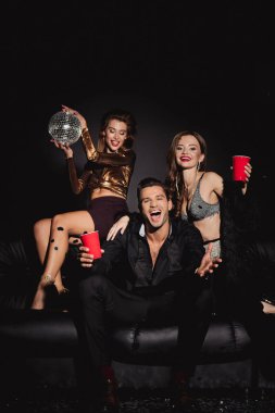 çekici ve gülümseyen arkadaşlar ellerinde disko topu ve siyah arka planda plastik bardaklar 