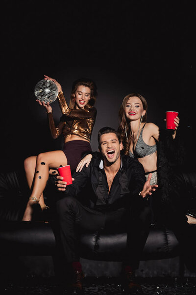 attractive and smiling friends holding disco ball and plastic cups on black background 