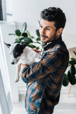 Gülümseyen iki ırklı adam Jack Russell Terrier 'ı tutuyor ve pencereden bakıyor. 