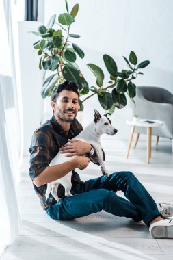Gülümseyen ve yakışıklı çift ırklı adam Jack Russell Terrier 'ı tutuyor.