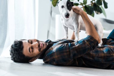 Gülümseyen ve elinde Jack Russell Terrier tutan iki ırklı yakışıklı bir adam.