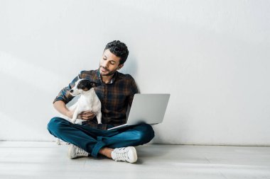 Dizüstü bilgisayarı olan yakışıklı, çift ırklı adam Jack Russell Terrier 'ı tutuyor.