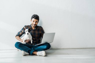 Yakışıklı ve gülümseyen çift ırklı dizüstü bilgisayarlı adam Jack Russell Terrier 'ı tutuyor.