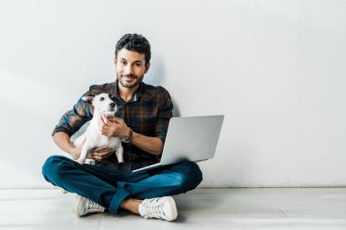 Yakışıklı ve gülümseyen çift ırklı dizüstü bilgisayarlı adam Jack Russell Terrier 'ı tutuyor.