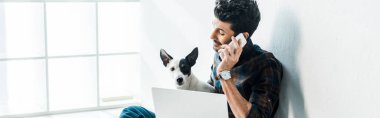 Akıllı telefondan konuşan ve Jack Russell Terrier 'ı tutan yakışıklı, çift ırklı adamın panoramik görüntüsü.