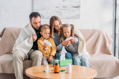 Hastalıklı bir aile, battaniyeye sarılmış, ilaçlarla birlikte masanın yanında oturuyor.