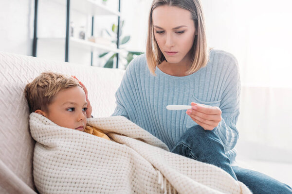 беспокойная женщина смотрит на термометр, сидя рядом с больным сыном
