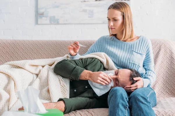 Uważna Żona Patrząc Termometr Pobliżu Chorego Męża Kichającego Leżąc Kolanach — Zdjęcie stockowe