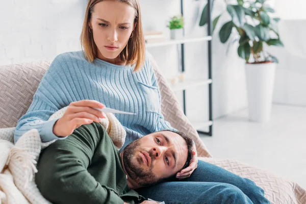 attentive wife looking at thermometer near diseased husband lying on her laps