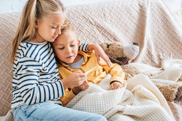 attentive sister looking at thermometer while hugging diseased brother