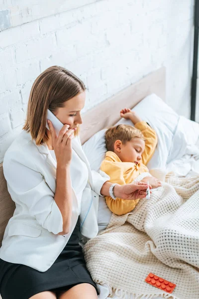 Worried Mother Looking Thermometer Talking Smartphone While Sitting Diseased Son — Stock Photo, Image
