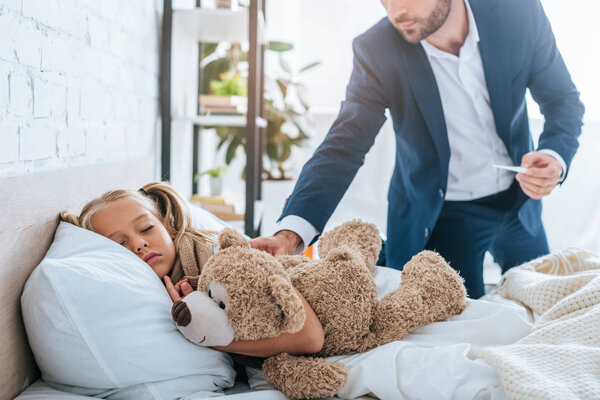 cropped view of father touching sick, sleeping daughter while holding thermometer