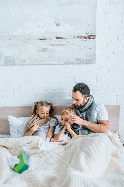 diseased man and children in warm scarfs lying in bed under blanket