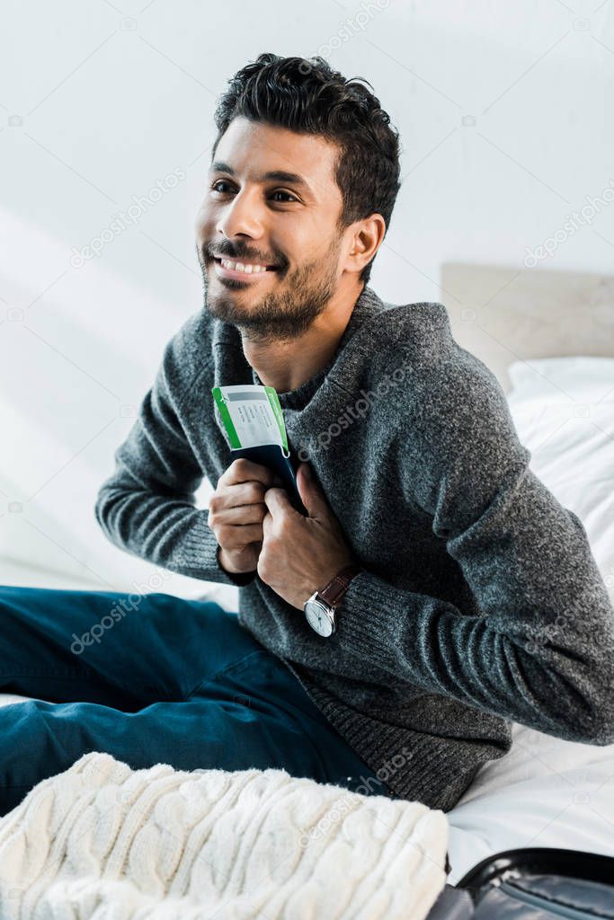 handsome and smiling bi-racial man in sweater holding passport with air ticket 