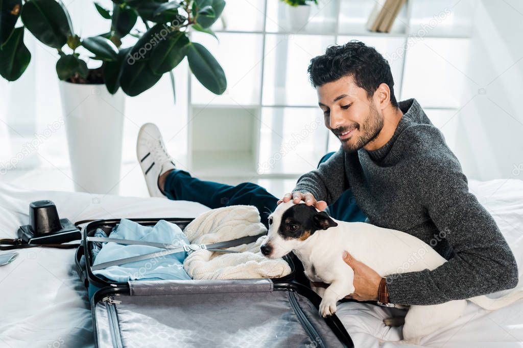 handsome and smiling bi-racial man stroking jack russell terrier and sitting near suitcase