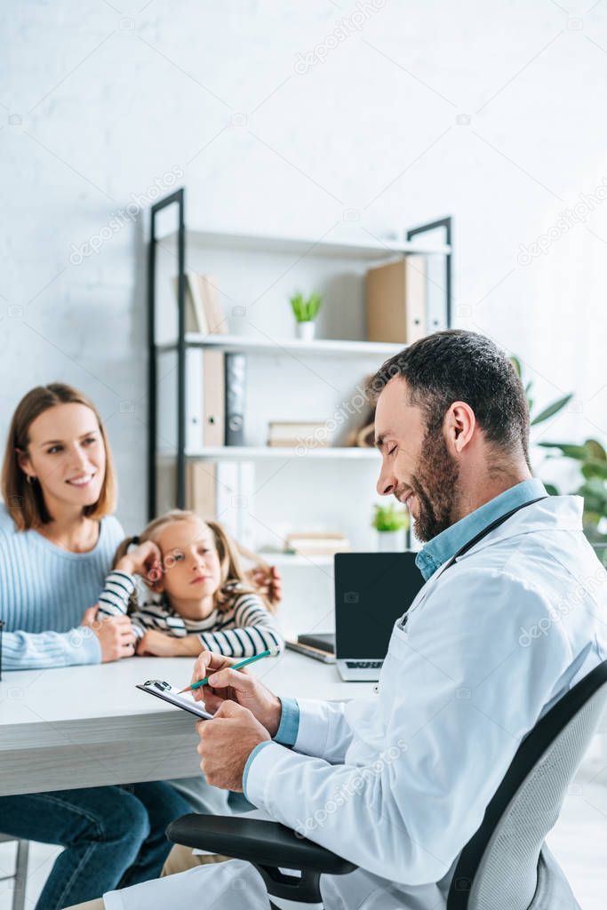 positive doctor writing prescription near smiling mother and daughter
