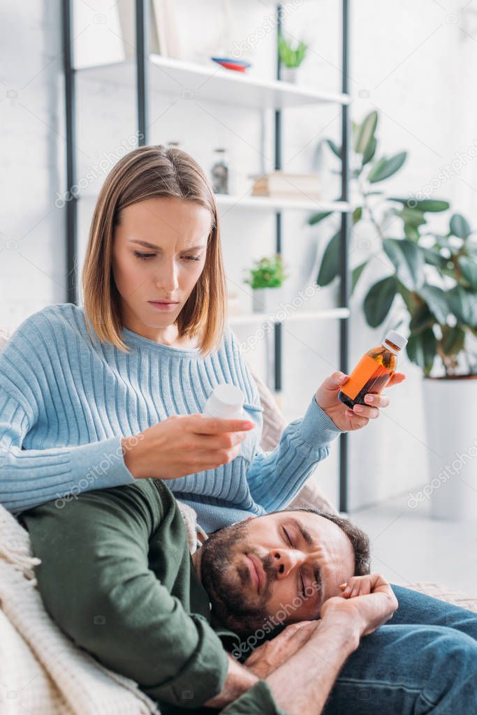 attentive wife holding container with pills and cough syrup near diseased husband lying on her laps