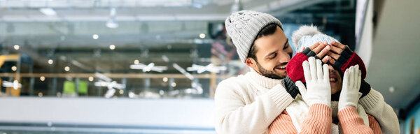 panoramic shot of handsome cheerful man closing eyes to woman to make a surprise on skating rink