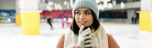 panoramic shot of happy woman in gloves and hat holding coffee to go on skating rink