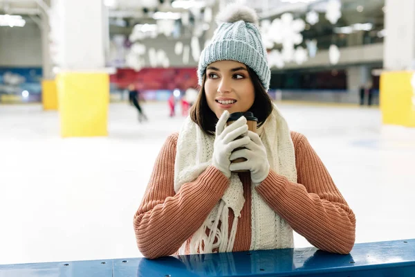 smiling woman in scarf, gloves and hat holding coffee to go on skating rink