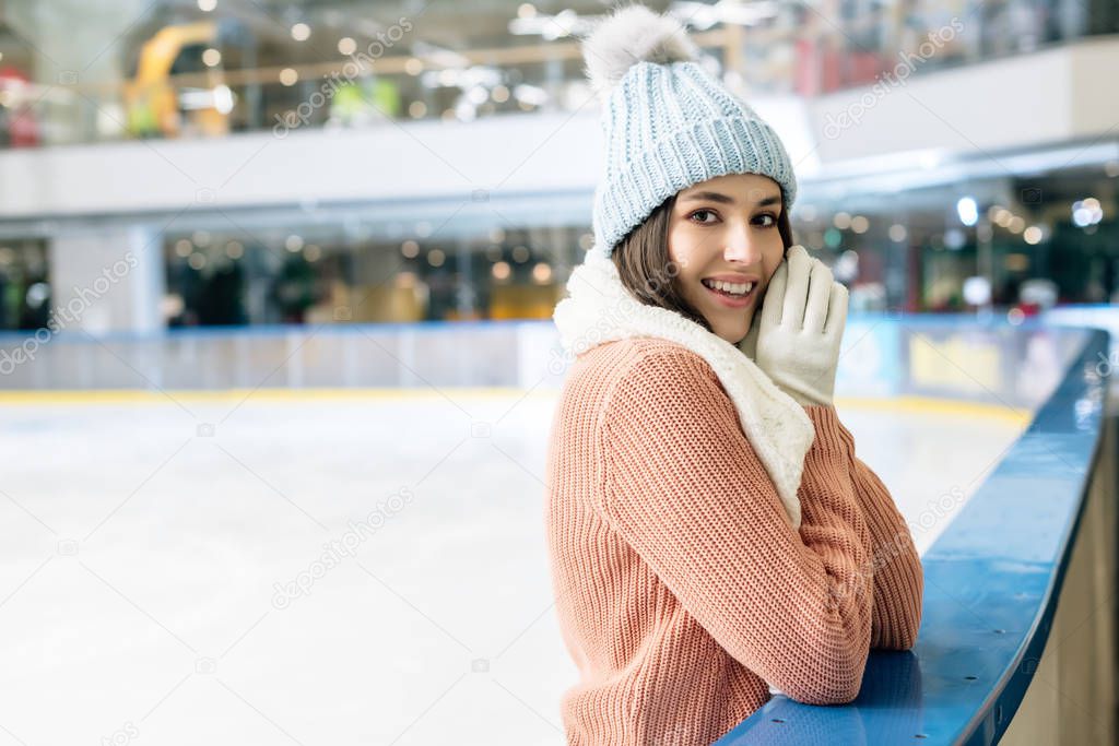 positive girl in sweater, scarf, gloves and hat standing on skating rink