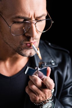 Handsome man in sunglasses lighting cigarette with lighter isolated on black clipart